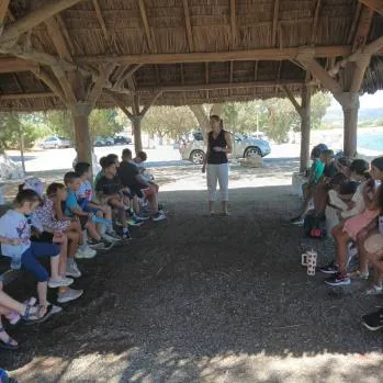 A facilitator speaking to participants seated under a wooden shelter